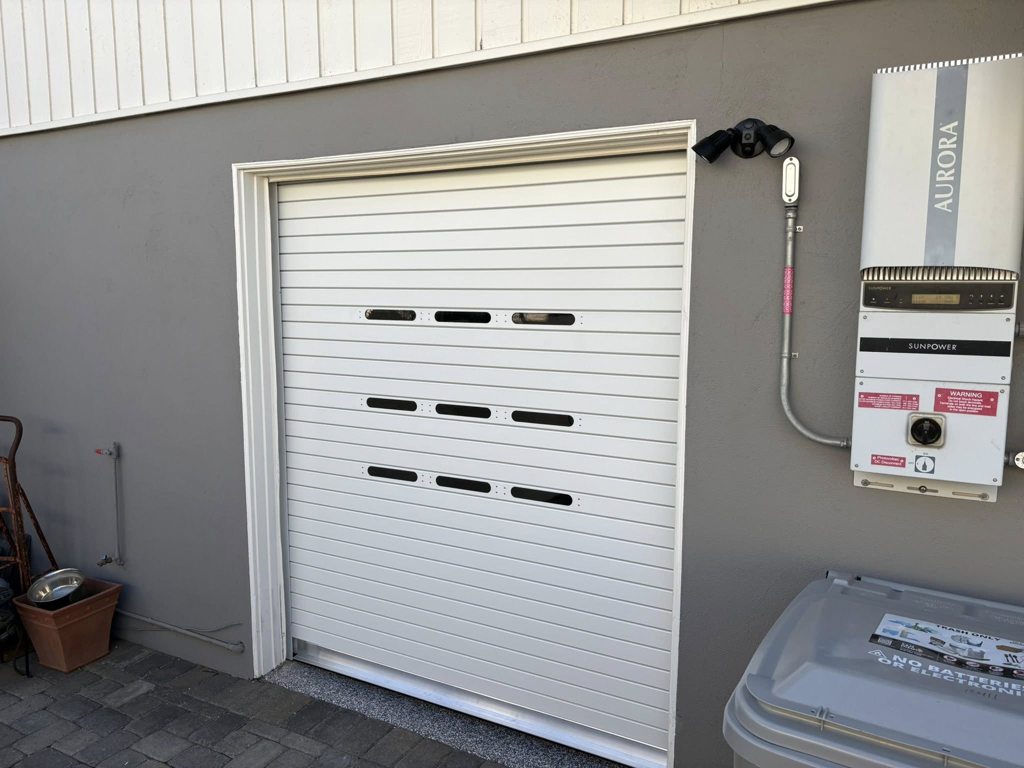 White garage door with horizontal vents, flanked by a utility box labeled Aurora, an outdoor camera above, and a trash bin nearby; brick paving in front.
