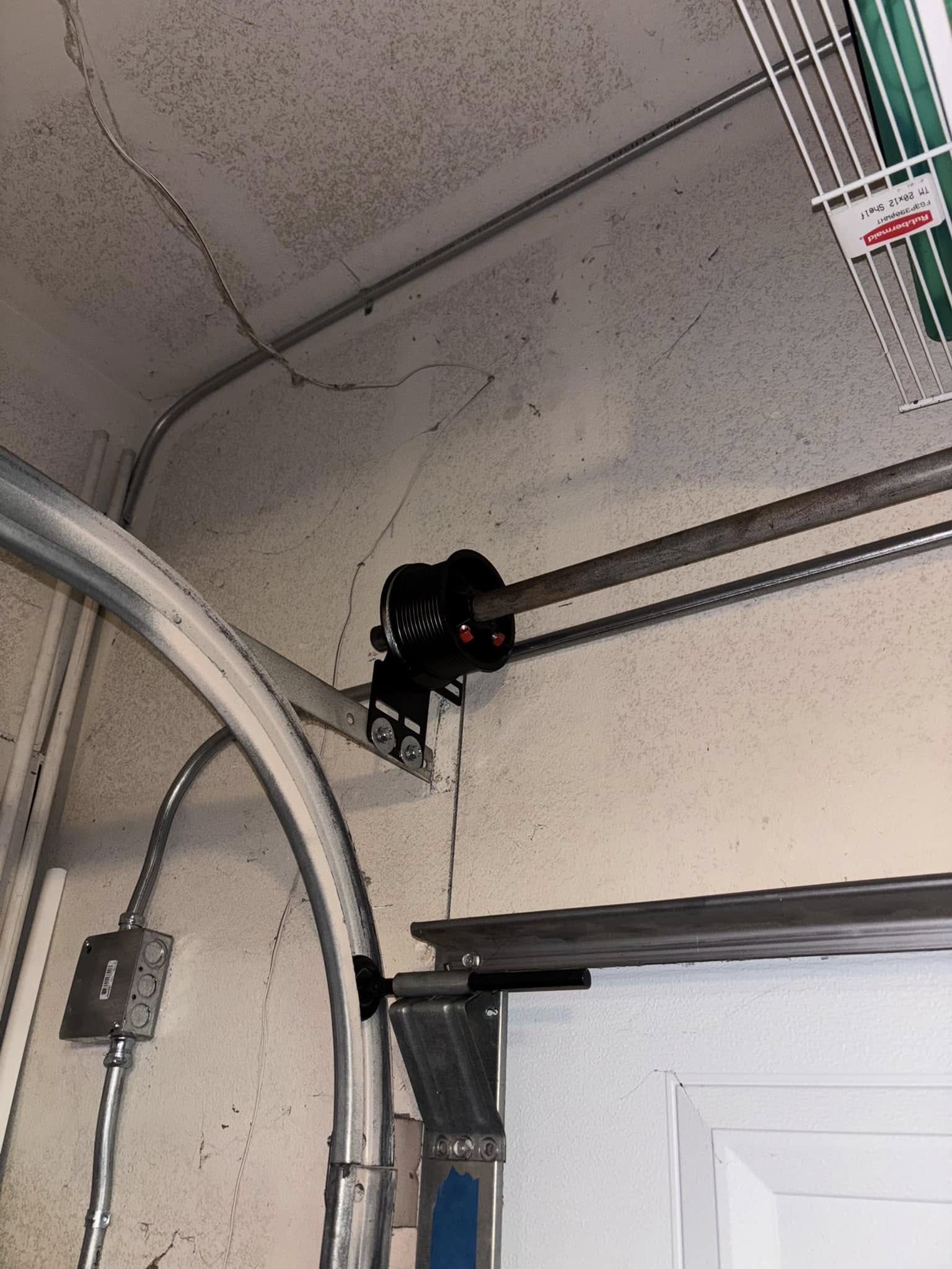 A black garage door cable spool mounted on the wall near the ceiling, with a metal rod and cable attached, next to electrical conduit and a white door.
