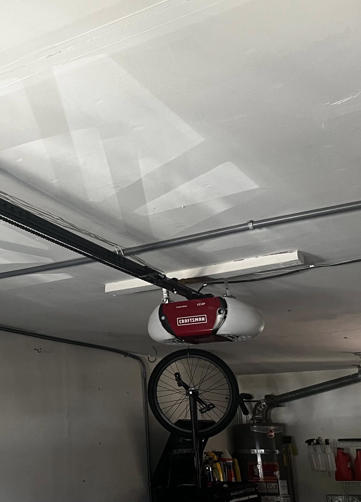 A red Craftsman garage door opener is mounted on the ceiling, with a bicycle wheel hanging nearby and shelves with various items visible in the background.