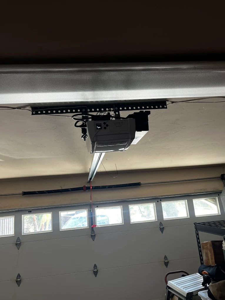 A garage door opener is mounted on the ceiling of a garage with a closed sectional garage door and windows letting in sunlight.