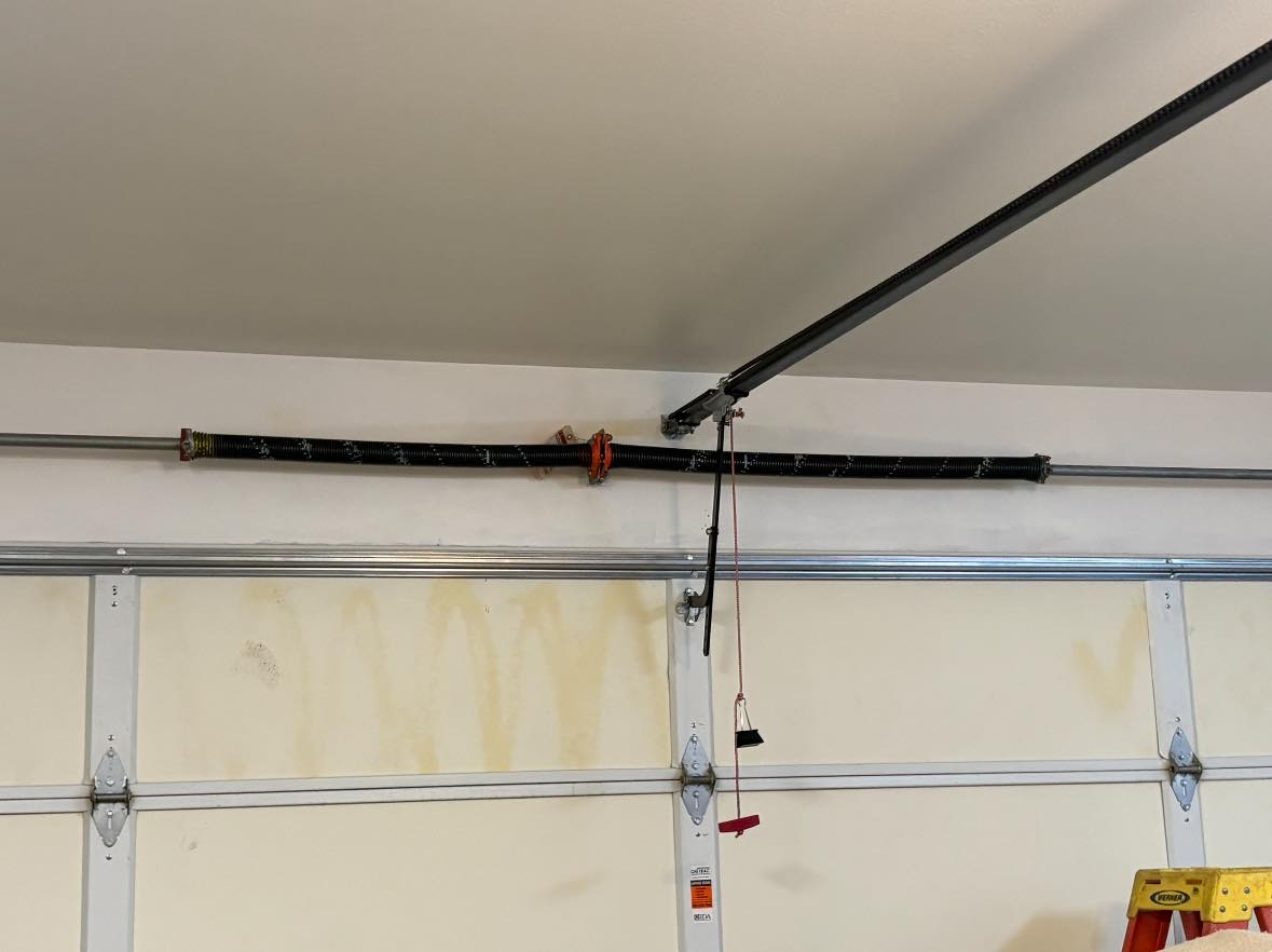 A garage door torsion spring with a winding bar and safety cable is mounted above a double garage door; a red cord hangs from the opener track.