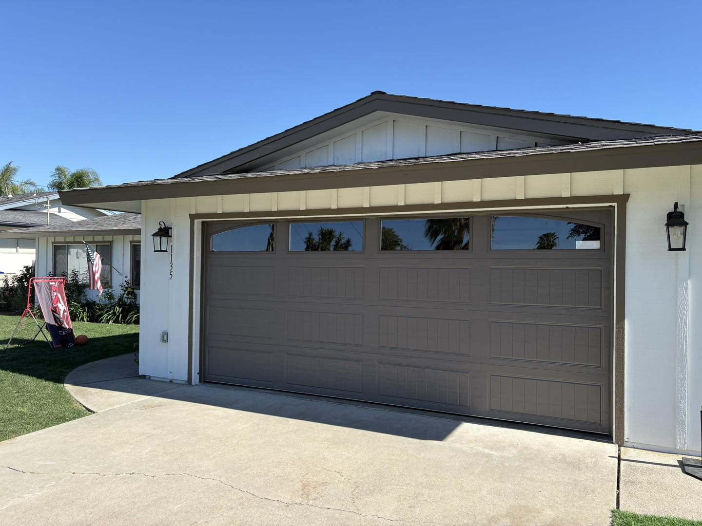 A single-story house with white siding and a brown garage door, flanked by two wall lanterns, with a concrete driveway and a lawn.