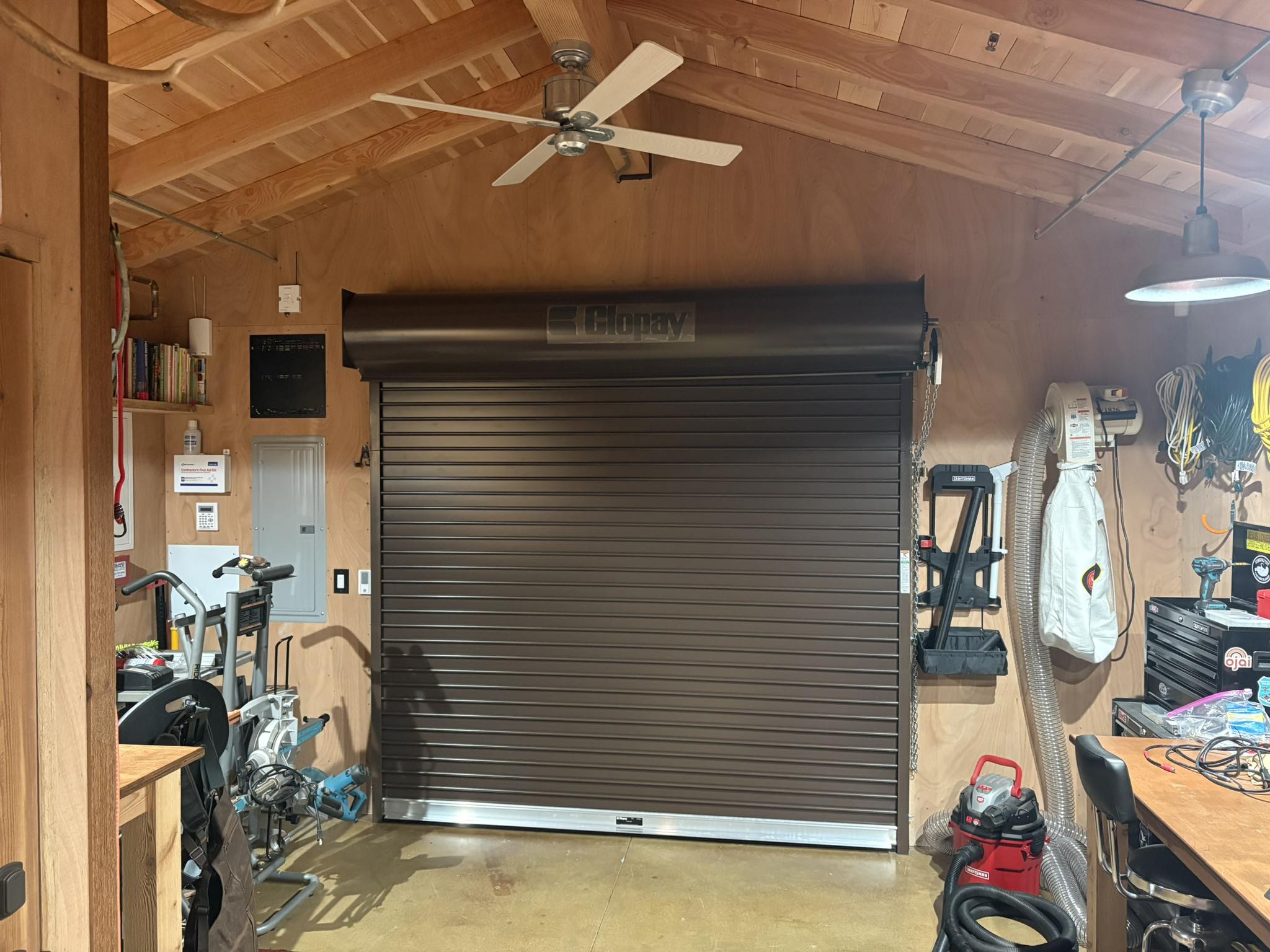 A closed brown roll-up garage door inside a well-lit wooden workshop, with various tools and equipment organized on either side.